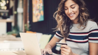 Eine junge Frau ist vor dem Laptop und benutzt ihre Debit- oder Kreditkarte fuer das Online-Shopping.